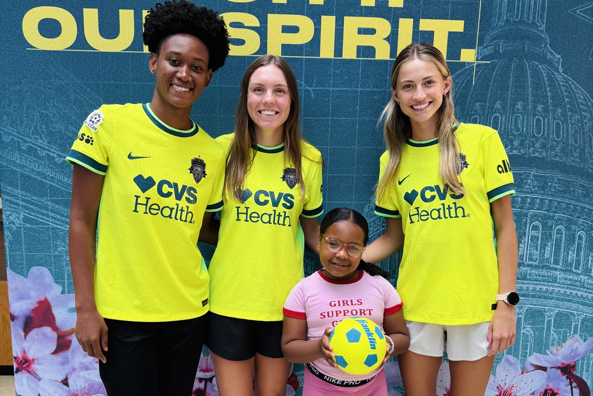 Three Washington Spirit players pose for a photo with a young girl holding a soccer ball at a visit to Children's National Hospital.