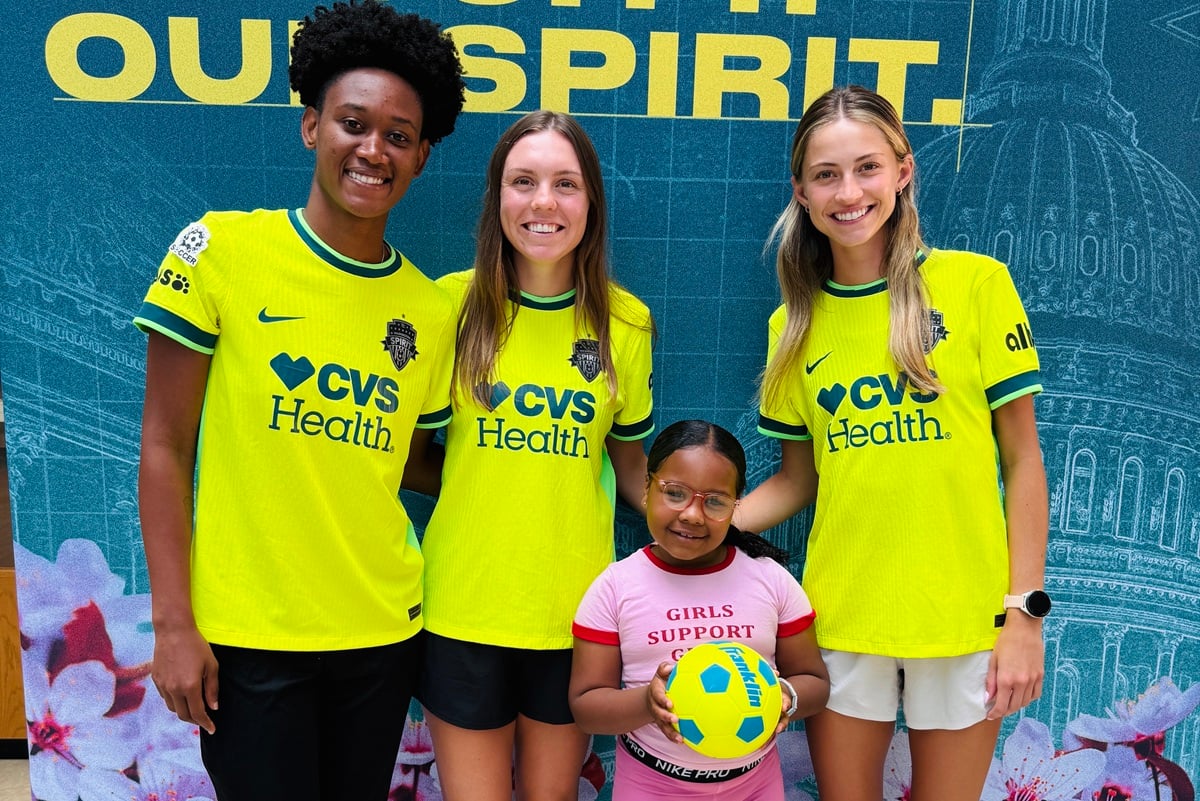 Three Washington Spirit players pose for a photo with a young girl holding a soccer ball at a visit to Children's National Hospital.