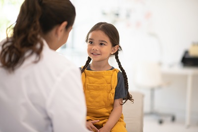 young girl wearing yellow overalls speaks with a provider in a white coat in the doctor's office