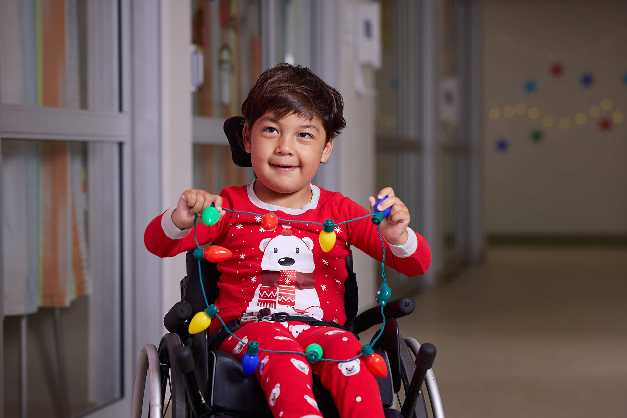 CNH patient Uly, age 4, sits in a wheelchair and handles a holiday light necklace while wearing winter-themed pajamas.